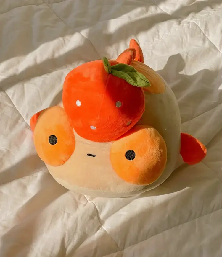 A plush orange goldfish with a tangerine on it's head, sitting in a sunbeam on a white quilt. 