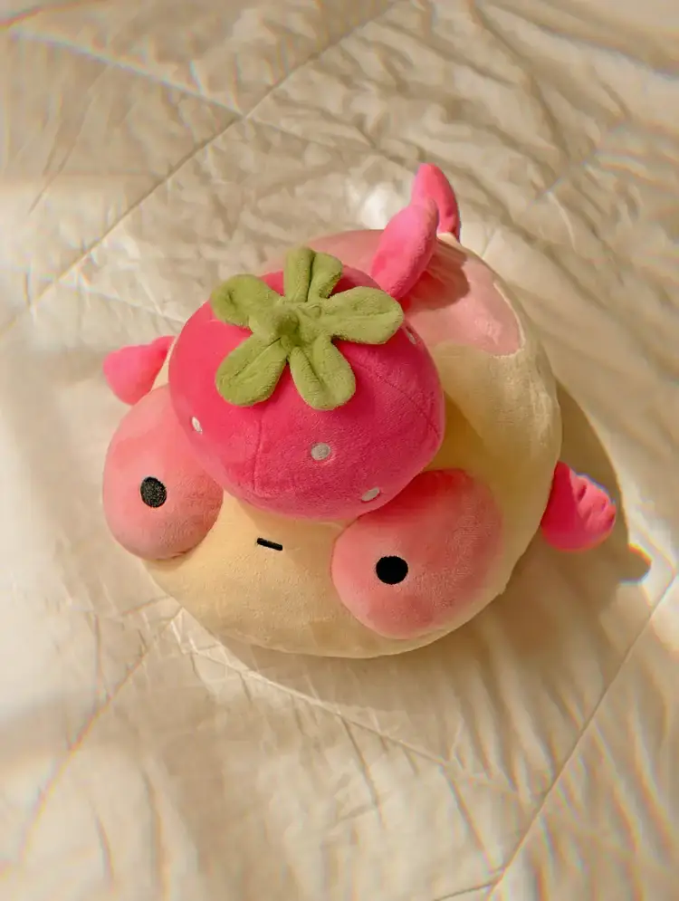 A plush pink goldfish with a strawberry on it's head, sitting on a white quilt. 